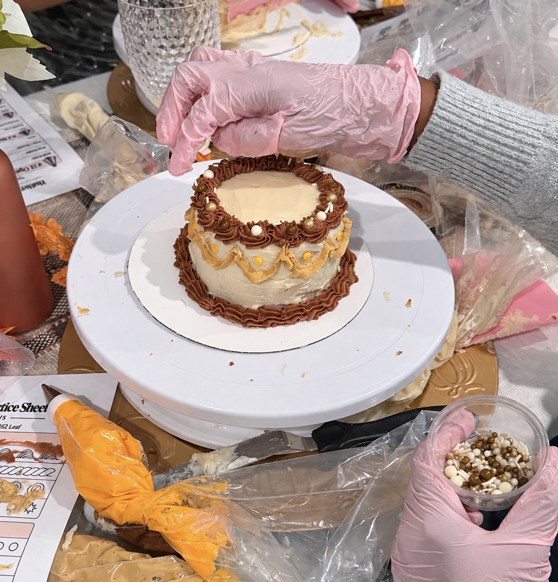 A guest finishing a small autumn-themed buttercream cake with chocolate drizzle borders and gold and white sprinkles